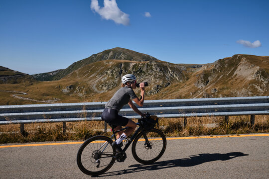 Woman cyclist is riding in the mountains and drinking water during her training with amazing view. Female cyclist in cycling kit and a helmet drinks water from a sports bottle.Transalpina, Romania