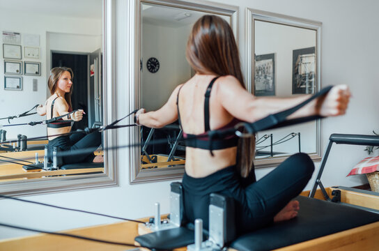 Fit-shaped female doing back strength exercises using pilates reformer machine in the sport athletic gym and looking at mirror wall. Active people training or medical rehabilitation concept image.