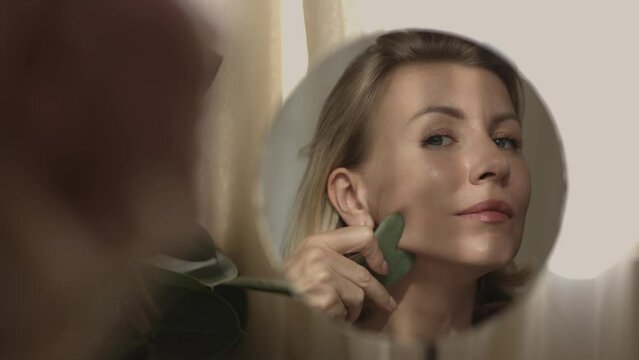 Middle aged beautiful woman making herself facial massage with gua sha behind the mirrow and watching herself in reflection at home