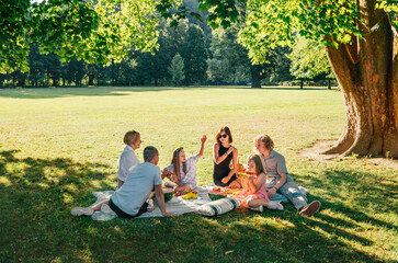 Big family sitting on picnic blanket in city park linden treeb during weekend Sunday sunny day. They are chatting and eating boiled corn and watermelon. Family values and outdoors activities concept