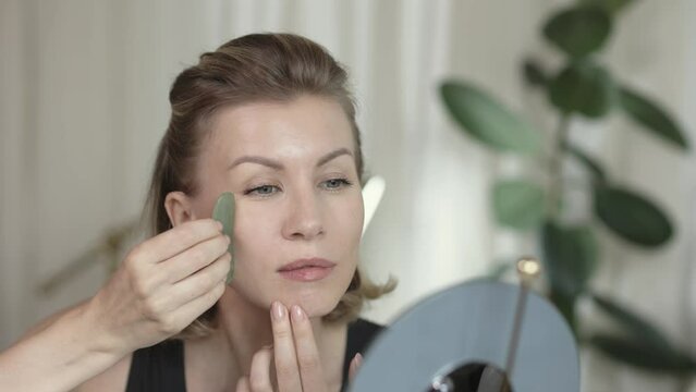 Middle aged beautiful woman making herself facial massage with gua sha behind the mirrow at home