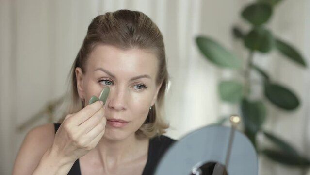 Middle aged beautiful woman making herself facial massage with gua sha behind the mirrow at home