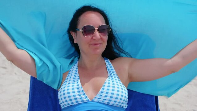 Happy woman having fun with scarf while relaxing on lounge chair. Female wearing sunglasses is enjoying during summer vacation at windy beach. She is spending leisure time on windy day.