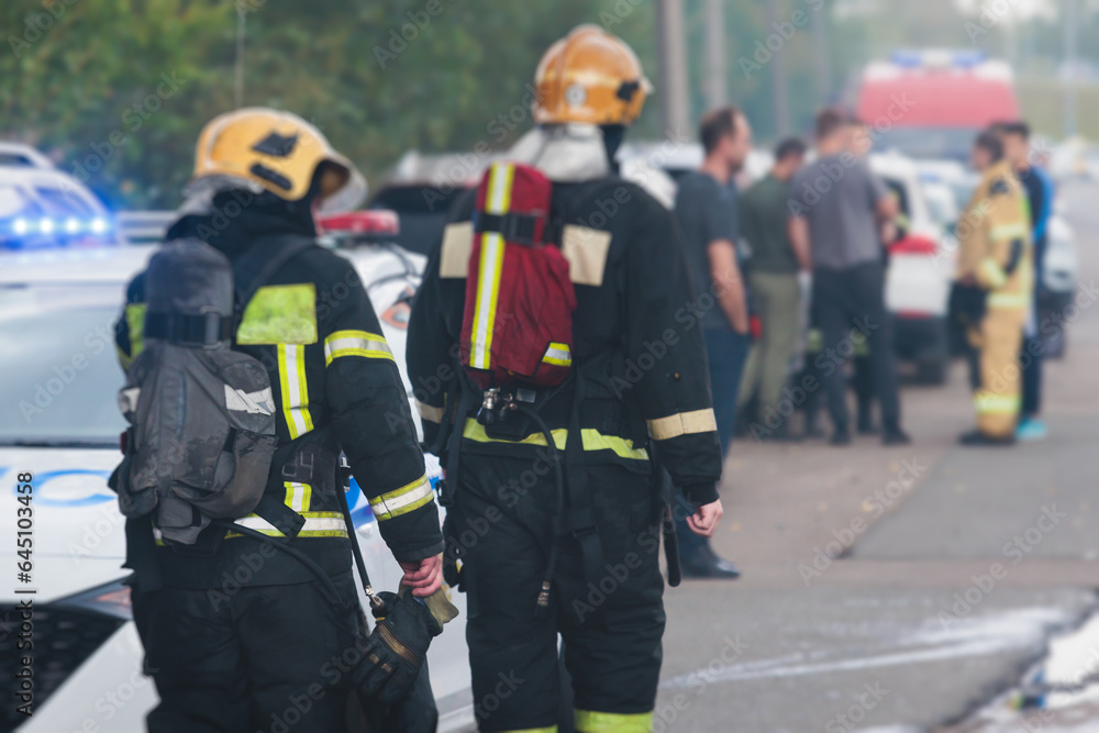 Wall mural Group of fire men in protective uniform during fire fighting operation in the city streets, firefighters brigade with the fire engine truck vehicle in the background, emergency and rescue service