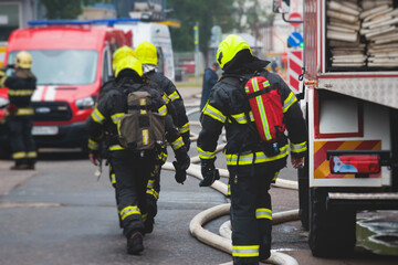 Obraz premium Group of fire men in protective uniform during fire fighting operation in the city streets, firefighters brigade with the fire engine truck vehicle in the background, emergency and rescue service