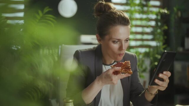 Sustainable workplace. happy modern 40 years old small business owner woman in a grey business suit in modern green office with pizza using smartphone applications.