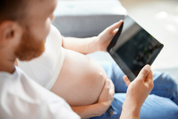 Cropped image of couple, future parents sitting at home and looking on tablet. Expecting baby, learning information. Concept of pregnancy, family, love, relationship, parenthood and childhood