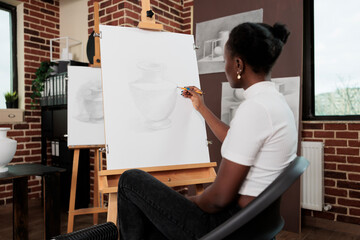 Young African American woman sitting at easel drawing vase on canvas while attending drawing classes, student girl improving sketching skills while studying in art school. Creative hobby concept