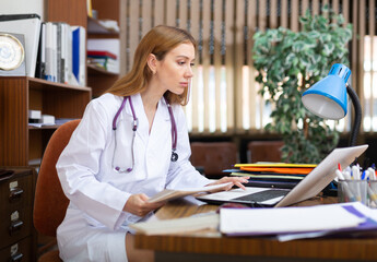 Young woman professional physician filling up medical forms on laptop while sitting at table in...