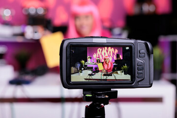 Asian vlogger holding notebook presenting product in front of camera while recording school supplies review using broadcast equipment. Content creator with pink hair live streaming