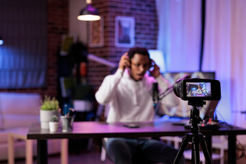 Focus shot on modern camera recording vlogger in blurry background talking about international news and events subjects. Man filming internet show using professional gadgets in rgb lights studio