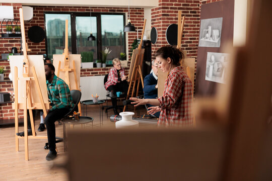 Group of different aged people sitting at easels studying drawing at group art class. Young woman teacher explaining still life sketching techniques to students. Social activities for adults