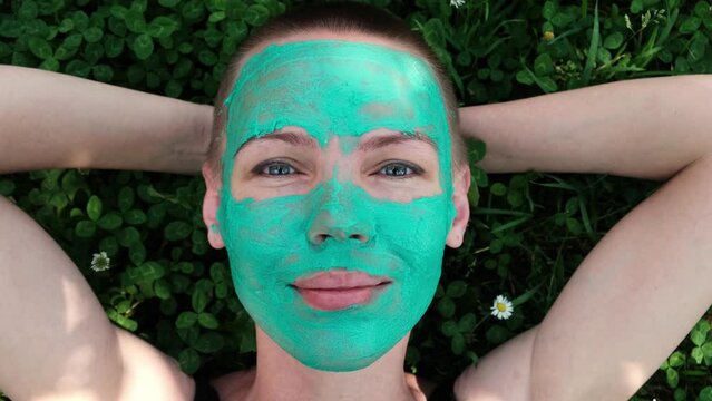 Woman with green cosmetic mask on her face is lying on grass.
