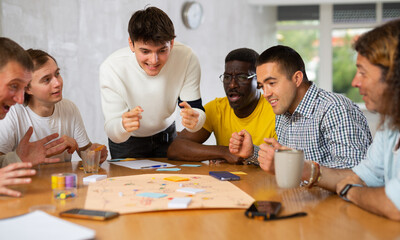 Excited man standing on foot playing board game with interested guys sitting around the table