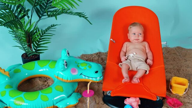 Tiny infant blond boy wearing shorts lies in the orange baby chair. Little kid on a beach vacation.