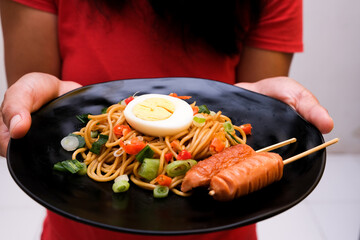 Noodles, eggs and sausages are served on a black plate, held by a woman's hand