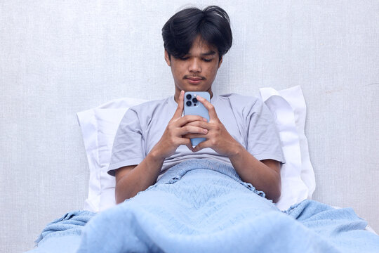 Calm young man in grey t-shirt lsit in bed reading message to check social media on smart phone at bedroom. 