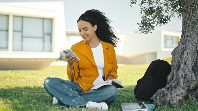 Full-length photo of a pretty, stylish positive brazilian or hispanic female student, sitting on the grass near the tree, using mobile phone, texting in social media with friends, smiling happily
