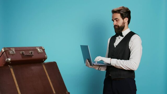 Hotel staff reviews list of guests on online bookings while using laptop and standing next to stack of suitcases. Employee working as doorkeeper providing an enjoyable interaction.