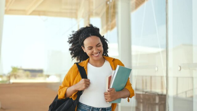 Happy lovely curly haired brazilian or hispanic female student, with a backpack, hold books and notebooks in her hand, walking near the university campus, looks away and smile, finished school day