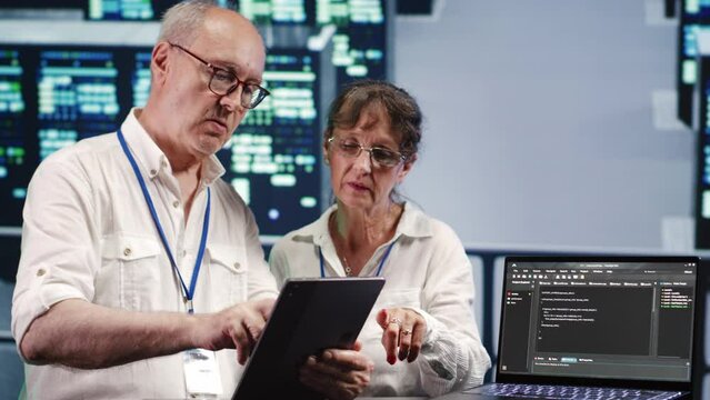 Data storage company executives solving tasks on tablet while running scripts on laptop terminal. Proficient tech support workers amidst servers, accessing databases and manipulating lines of code