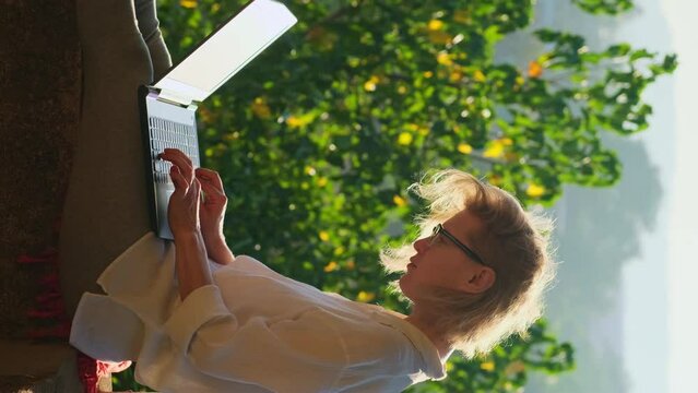Vertical shot of blonde woman typing on a laptop keyboard in sunshine outdoor. Good feeling female writer working on the rooftop with a beautiful park view at sunrise.