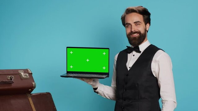 Classy bellboy shows greenscreen on laptop in studio while he wears white gloves next to guests suitcase. Luxurious stylish doorman working at modern hotel, presenting isolated mockup display.