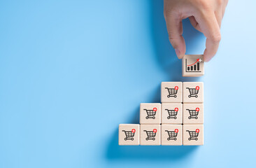 Hand holding wooden cube block stacking with investment increasing trend-up graph and shopping trolley cart. Increase higher sale volume for online e-commerce business marketing strategy concept.