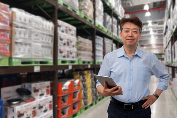 a middle-aged Asian businessman holding a tablet at a warehouse full of goods