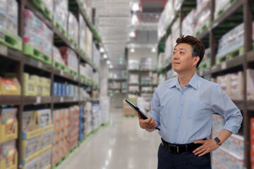 a middle-aged Asian businessman holding a tablet at a warehouse full of goods