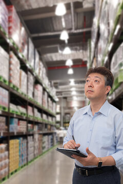 a middle-aged Asian businessman holding a tablet at a warehouse full of goods