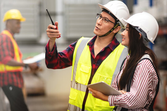 Father or Senior supervisor advises a young worker about warehouse work, both wore safety vest and hardhat for safety, pointing with  walkie-talkie