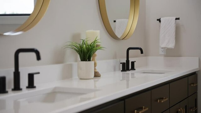 Tight panning shot of a white bathroom countertop with 2 black faucets and a green plant