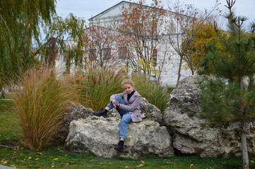 beautiful girl in a plaid coat on a walk in the autumn park posing near a large stone. the child smiles and looks a little to the side. active lifestyle and outdoor activities. attractive people
