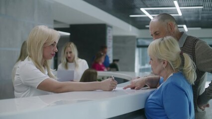Elderly couple stand near reception desk in clinic lobby area and make appointment with doctor. Female administrator talks with patients at information counter. Medical staff work in modern hospital.