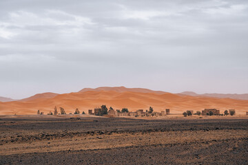 Fototapeta na wymiar Houses and palm trees in front of the big Sahara Desert in Merzouga, Morocco
