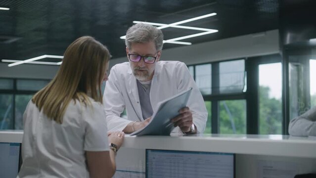 Mature doctor stands near reception desk in clinic waiting room and discusses work with nurse. Female administrator talks with colleague at information counter. Medical staff work in modern hospital.