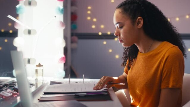 Teenage girl at home in bedroom studying online with laptop and making notes in folder- shot in slow motion
