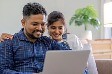 Portrait of happy Indian couple using laptop together while sitting on sofa in living room at home.