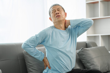 Fototapeta premium Elderly Asian man on sofa, grimacing from spinal pain, seeking relief from osteoporosis-related discomfort at home.