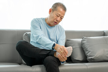 Senior Elderly Asian man in blue shirt suffering from intense foot and ankle pain on the sofa.