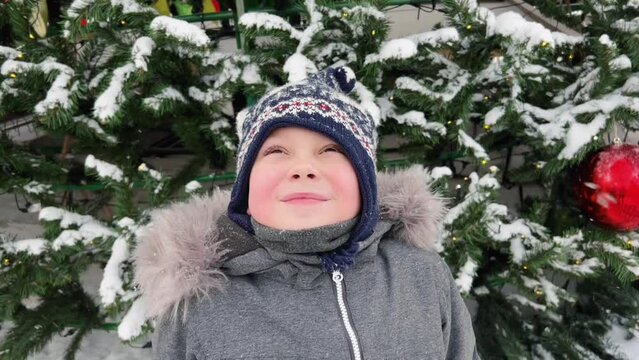 winter video portrait of cute caucasian boy wearing scandinavian hat standing by christmas tree looking at snow faling down slowly. 