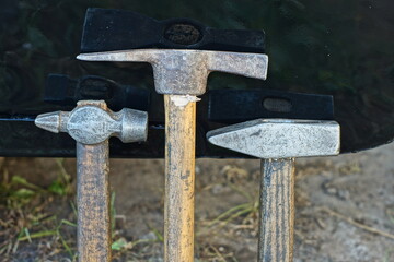 three old iron mechanical hand industrial heavy retro hammers with wooden handles stand on the ground near a black iron painted surface on the street during the day