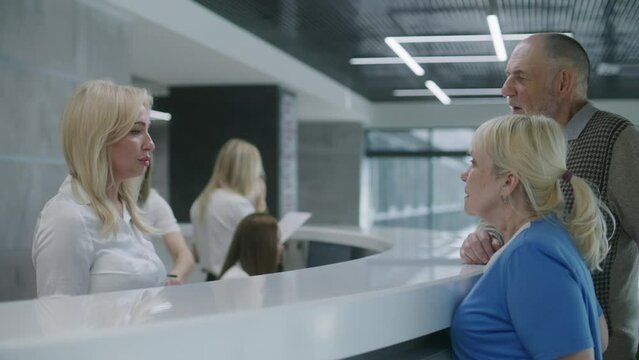 Elderly couple come to reception desk in clinic lobby entrance and make appointment with doctor. Female administrator talks with patients at information counter. Medical staff work in modern hospital.