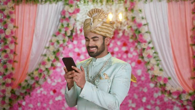 Happy smiling young man as Groom or wedding ethnic wear using mobil phone on decorated background - concept of social media sharing, technology and conversation