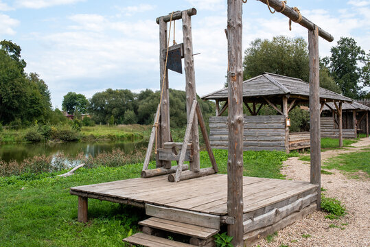 A guillotine on a wooden platform, behind it a river and a wooden barn.