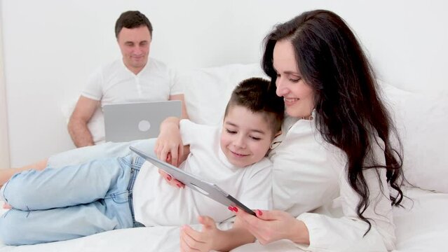 freelance work allows family to be together father works on computer laptop in bed at home mom and son play on tablet have great time with family woman and boy have fun relaxing man earn money