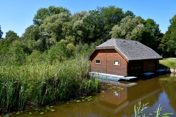 A view of a small house, shack, or shelter with an angled roof made out of thatch located next to a shallow pond or river overgrown with reeds, grass, and other flora seen in Poland in summer