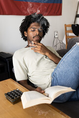 Afro boy with curly hair smoking on the sofa at home while picking up a book. Vintage vertical photo