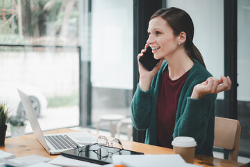 Attractive young woman talking on the mobile phone and smiling while sitting at her working place in office and looking at laptop